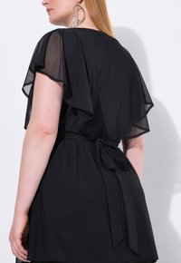 Abito nero con maniche trasparenti a fluttuare, vita legata e parte inferiore ampia. Struttura liscia con elementi di design minimi. Orecchini a cerchio d'argento.