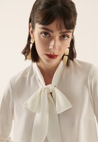 White blouse with a tied bow detail at the collar. Model wears large beige geometric earrings. Smooth texture with minimal design elements.