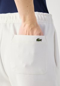 Baltas teksturēts audums bikses ar aizmugures kabatu, kurā ir zaļš izšūts krokodila logo. Augšpusē redzama elastīga josta.