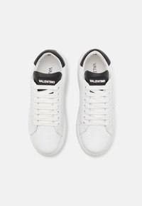 White leather sneakers with black accents, featuring a rounded toe, lace-up closure, and brand logo on the tongue. Smooth surface texture.