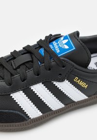 Baskets en cuir noir avec trois rayures blanches et le texte doré "SAMBA" sur le côté. Présente une étiquette bleue et des lacets noirs. Semelle en caoutchouc texturée.