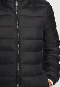 Veste en duvet noire avec des panneaux matelassés horizontaux, une fermeture éclair à l'avant et des poignets élastiques pour une meilleure isolation et confort.