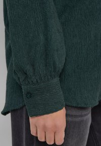 Close-up van een hand die een donkergroene gestructureerde lange mouw shirt met een geknoopte manchette over donkergrijze broeken draagt.