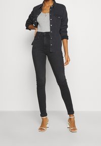 Jeans skinny de tiro alto en negro combinados con una camiseta sin mangas ajustada en gris y una camisa de mezclilla oscura. Acabados con unos tacones blancos de tiras.
