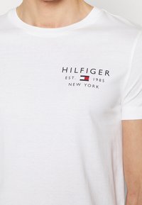 Bílý bavlněný tričko s černým logem a červenými a modrými akcenty, na přední straně s textem "HILFIGER EST. 1985 NEW YORK".