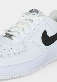 Λευκό αθλητικό παπούτσι Nike με μαύρο λογότυπο swoosh, λευκά κορδόνια περασμένα μέσα από μεταλλικό ταμπελάκι κορδονιών και κείμενο "NIKE EST. 1972" στη γλώσσα.
