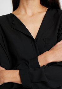 Blusa nera con scollo a V, maniche lunghe con polsini abbottonati e taschino frontale; tessuto leggero e liscio con una leggera lucentezza.