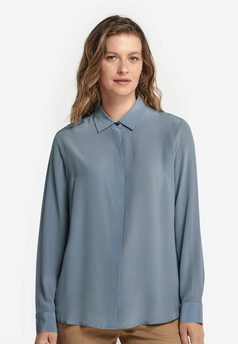 Blusa azzurro chiaro a maniche lunghe con colletto, texture liscia, leggero lucido e chiusura a bottoni sul davanti.