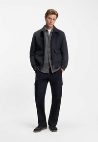 Navy jacket with a front zip, two patch pockets, and ribbed cuffs. Layered over a gray button-up shirt and paired with dark cargo pants.