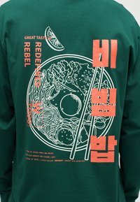 Zielona bluza z białym rysunkiem miski bibimbap, pałeczek, plasterka cytryny i czerwonym tekstem w języku angielskim i koreańskim.