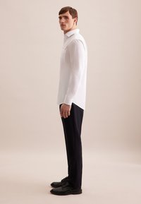 Camicia bianca a maniche lunghe con colletto a oblò e taschino, abbinata a pantaloni neri e scarpe stringate nere.