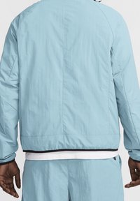 Ljusblå vindjacka med en slät yta, svart kant i nederkant, och en avslappnad passform, med raglanärmar och en enkel design.