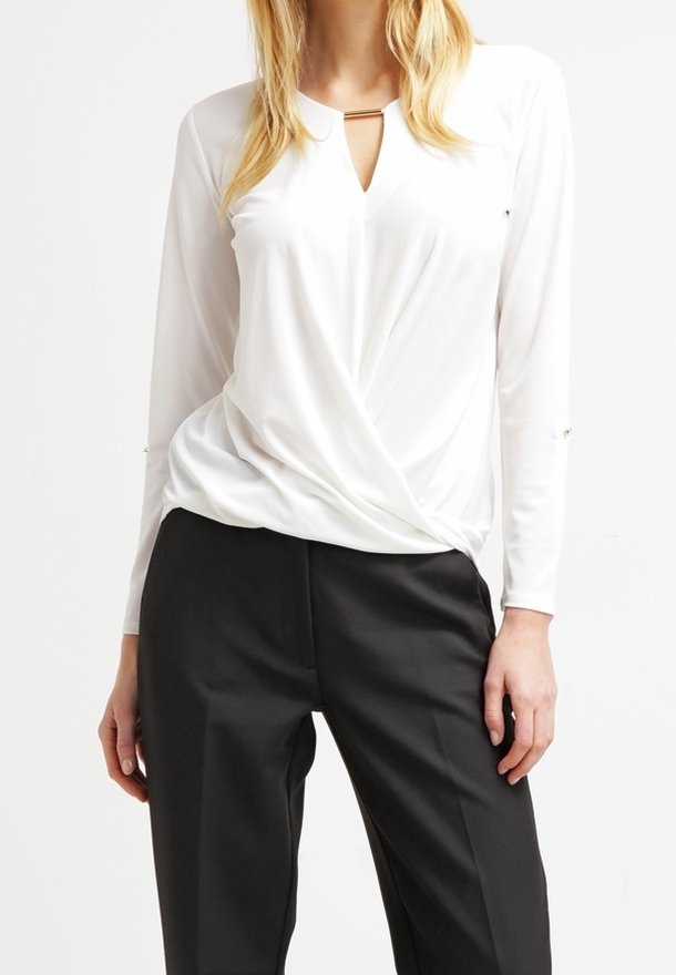 Blusa bianca a maniche lunghe con design a torsione frontale, caratterizzata da un collo parziale a forma di chiave e un tessuto morbido. Abbinata a pantaloni neri a gamba larga.