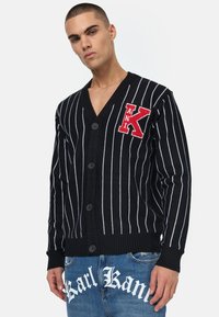 Svart cardigan med vertikale hvite striper, V-hals, fem knapper og broderte røde 'K' logo på venstre bryst. Strukturert strikket stoff.