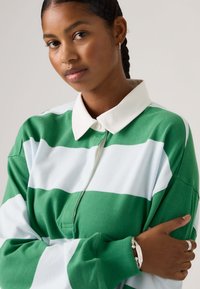 Νεαρή γυναίκα με πλεξούδες, που φοράει μακρυμάνικο πουκάμισο με πράσινες και λευκές ρίγες και λευκό γιακά, ασημένιο βραχιόλι και δαχτυλίδι, με τα χέρια σταυρωμένα.