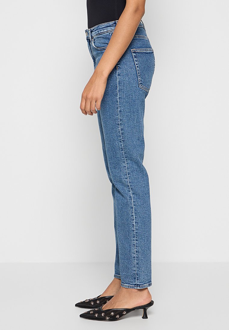 Helle blaue Denimjeans mit geradem Bein. Zu den Merkmalen gehören fünf Taschen, Gürtelschlaufen und dezente Nähte. Getragen mit schwarzen Mules.