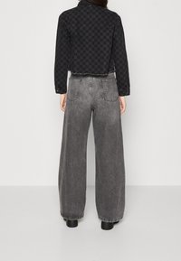 Woman wearing black and gray checkered cropped jacket with high-waisted, wide-leg faded gray jeans and black shoes, standing against white wall.