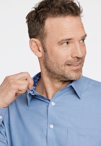 Ljusblå skjorta med en texturerad tyg, knäppt krage och blommönstrad foder synligt. Har en patchficka och vita knappar.