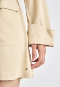 Vêtement beige au toucher lisse, avec un poignet replié et un petit emblème de marque à l'ourlet. Gros plan sur la main sur le tissu.