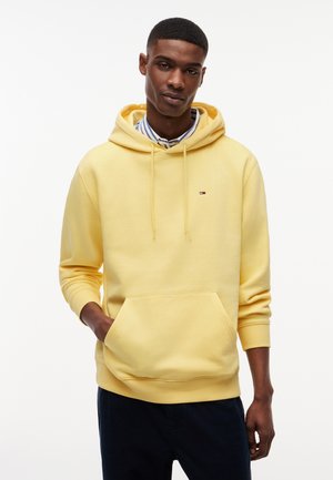 Bărbat tânăr purtând un hanorac galben deschis peste o cămașă cu guler, albastru cu dungi albe, cu o mână în buzunarul din față, stând în picioare pe un fundal alb.