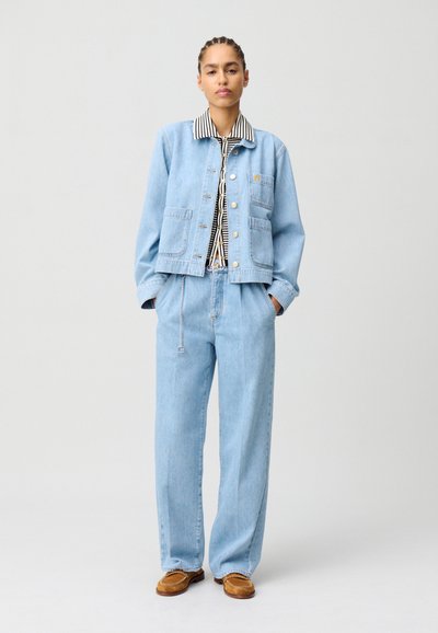 Veste en denim bleu clair avec un col rayé, dotée de deux poches avant et de boutons dorés, associée à un jean large assorti et des chaussures marron.