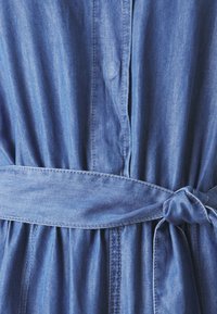 Robe en denim avec une taille à nouer, présentant une fermeture à boutons sur le devant, un tissu froncé et une couleur bleu moyen avec une texture subtile.