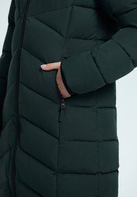Abrigo acolchado verde oscuro con un patrón de acolchado diagonal, bolsillo lateral con cremallera y textura de tela suave.