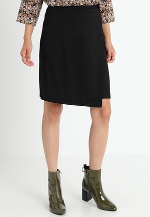 Schwarzer knielanger Rock aus glattem Stoff mit schräger Saumlinie. Kombiniert mit glänzenden olivgrünen Stiefeletten mit klobigem Absatz.