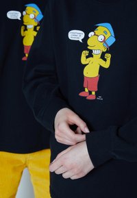 Mustapaita, jossa on etupuolella sarjakuvahahmo, jolla on keltainen vartalo, sininen hattu ja punaiset shortsit. Hihoissa on resorit.