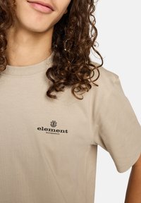 Camiseta de algodón beige de manga corta, con un logo impreso en negro que dice "element" en el pecho. Textura suave y corte casual.