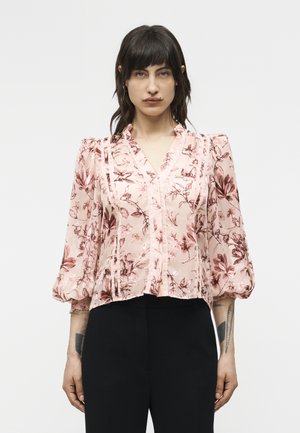 Kvinde iført lyserød blomstret bluse med puffede ærmer og sorte bukser, stående mod en ensfarvet hvid baggrund.