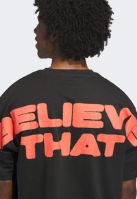 Black cotton t-shirt featuring bold, textured "BELIEVE THAT" text in bright coral on the back. Short sleeves, relaxed fit.