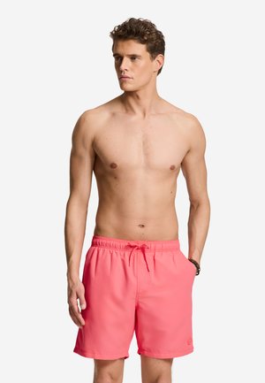 Bărbat fără tricou, purtând pantaloni scurți de baie roz coral cu șnur, stând în fața unui fundal alb simplu, privindu-se în partea laterală.