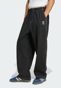 Black wide-leg pants with white vertical stripes, elastic waistband, side pockets, and logo patch. Paired with blue shoes featuring white accents.
