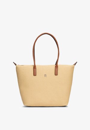 Beežist kootud kandekott pruunide nahast käepidemetega ja väike metalliklogo esiküljel keskel.