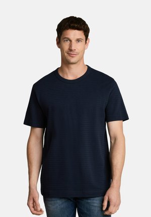 Man met kort bruin haar, draagt een effen donkerblauw T-shirt en blauwe spijkerbroek, staand tegen een witte achtergrond.