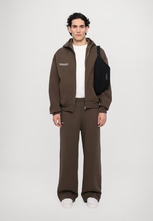 Bruine zip-up sweatshirt met capuchon, witte t-shirt eronder, bijpassende bruine sweatpants en een zwarte crossbody tas. Casual stof en losse pasvorm.