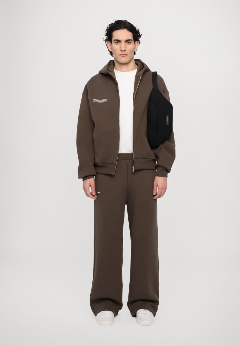 Rjav zip-up pulover s kapuco, pod njim bela majica, usklajeni rjavi trenirke in črna torba čez ramo. Ohlapen kroj in udoben material.