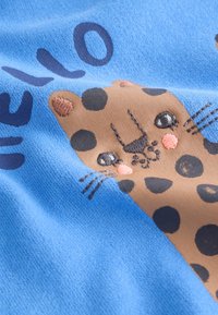 Tissu bleu avec un visage de chat marron tacheté brodé et le mot "HELLO" en lettres bleu foncé.