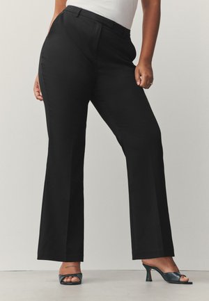 Gros plan d'une personne portant un pantalon noir taille haute à jambes larges et des sandales noires à talons ouvertes, debout devant un fond uni.