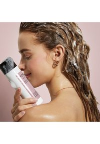 Vit schampoflaska med svart lock märkt "KIKO" och rosa detaljer. Personen har vått, vågigt hår med skum och bär guld-örhängen i ringar.