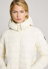 Švarkas pūkų puffer tipo, šviesios grietinėlės spalvos, su aukštu apykakle, horizontaliais siuvinėjimais ir priekiu užtraukiamu užtrauktuku. Juoda detalė ant rankovės.