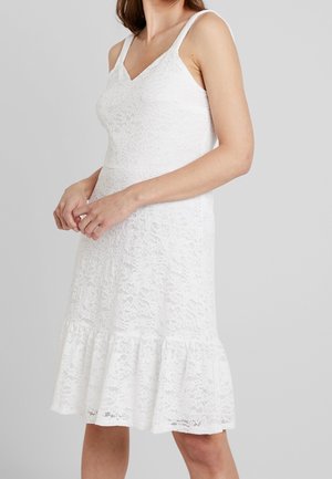 Femme portant une robe blanche en dentelle sans manches avec un ourlet à volants, debout devant un fond gris clair uni.