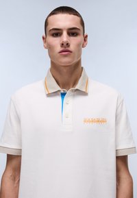 Beige polo-paita, jossa on teksturoitu kuvio, oransseja ja sinisiä yksityiskohtia kauluksessa sekä logo vasemmassa rinnassa.