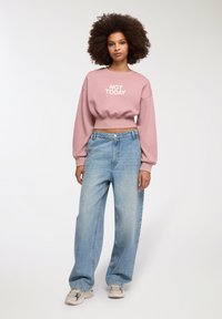 Pullover rosa corto con la scritta "NOT TODAY", abbinato a jeans a gamba larga blu chiaro e sneakers neutre. Tessuto morbido, vestibilità comoda.
