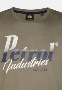 Olivově zelené bavlněné tričko s kulatým výstřihem, na kterém je lesklé logo s modro-bílým přechodem, na němž je nápis "Petrol Industries EST. 1989."