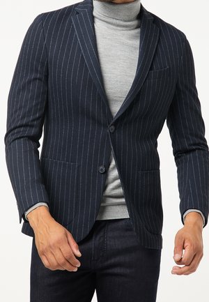Mann trägt einen dunkelblauen Nadelstreifenblazer über einem hellgrauen Rollkragenpullover und dunklen Hosen.