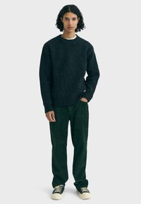 Suéter de punto oscuro con textura y canalé, combinado con pantalones de pana verdes. Los zapatos son negros con acentos blancos. Cuello redondo simple.