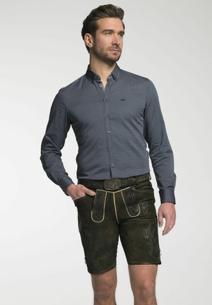 Herrenoutfit mit einem navyblauen Polka-Dot-Hemd und braunen Wildleder-Lederhosen, die mit geprägten Details und einem kontrastierenden goldenen Gürtelbesatz versehen sind.