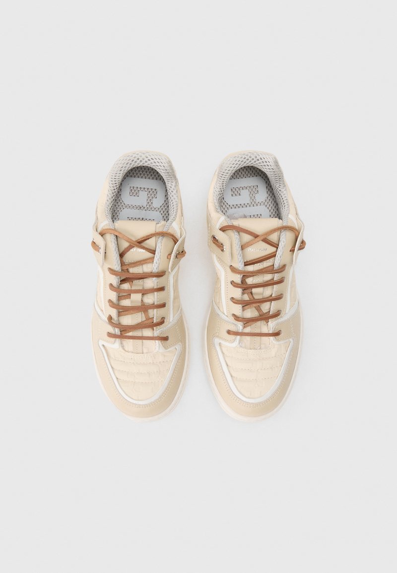 Beige sneaker with textured upper, brown laces, and white accent stitching. Mesh tongue and cushioned interior. Decorative branding visible.